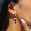 Close-up of an ear wearing a gold earring with a pearl drop, with a hand touching the skin.