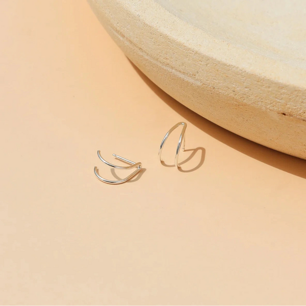 A pair of thin silver wishbone-shaped stud huggie earrings resting on a warm peach surface next to a textured beige ceramic plate.