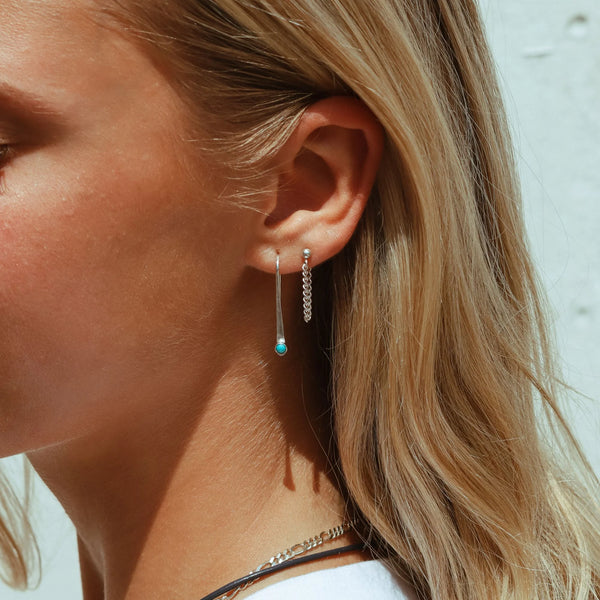 A close-up side profile of a woman with blonde hair wearing a pair of long silver dangle earrings, one with a small turquoise bead and one with a textured chain.