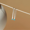 A pair of sterling silver arc-shaped drop earrings with a small turquoise gemstone at the end hanging from a wood skewer against a soft beige background.
