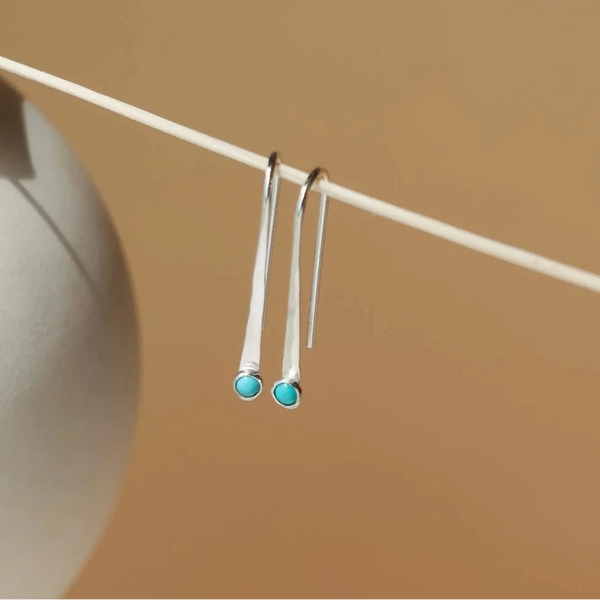 A pair of sterling silver arc-shaped drop earrings with a small turquoise gemstone at the end hanging from a wood skewer against a soft beige background.