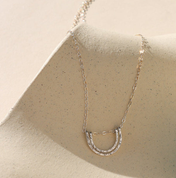 A silver chain necklace with a double U-shaped arch pendant on a shadowy neutral background.