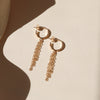 A pair of gold hoop earrings with three strands of chain fringe on a shadowy neutral background.