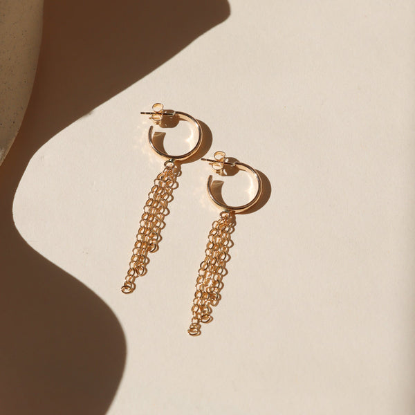 A pair of gold hoop earrings with three strands of chain fringe on a shadowy neutral background.