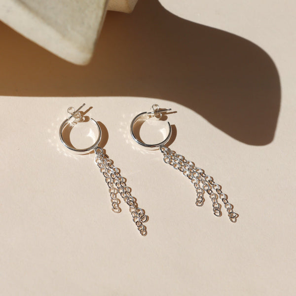 A pair of silver hoop earrings with chain fringe against a shadowy neutral background.