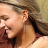 A person with dark blonde hair wearing a silver hoop earring with chain fringe standing outdoors.
