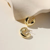 A pair of chunky gold hoop earrings against a soft beige stone backdrop.