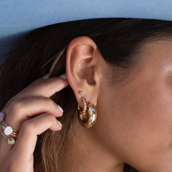 A person wearing a chunky gold hoop earring stacked with a smaller braided hoop and mini stud, their hand, covered in rings, is pulling their hair back.