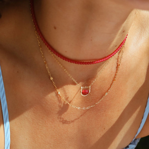 A close-up of a person's neck and chest wearing a layered necklace set. The set includes a red beaded choker, a delicate gold chain with a small oval ruby pendant, and a thin gold chain with tiny gold beads.