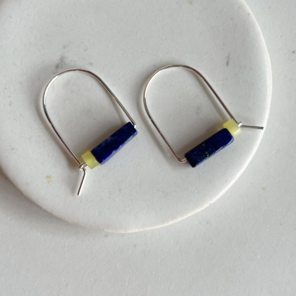 A pair of sterling silver arch earrings with a bar of lapis stone and a lemon jade bead laying against a marble surface. 