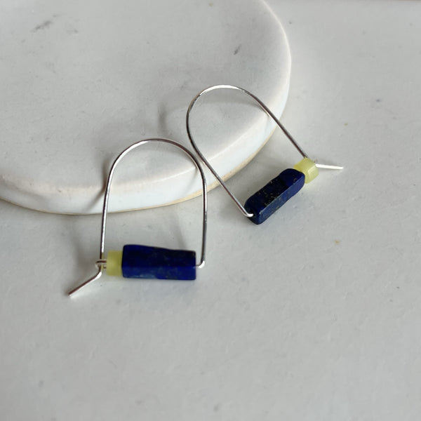 A pair of sterling silver arch earrings with a bar of lapis stone and a lemon jade bead leaning against a marble surface. 