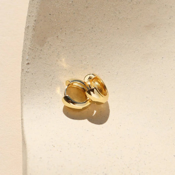 A pair of shiny gold huggie hoop earrings on a beige stone surface.