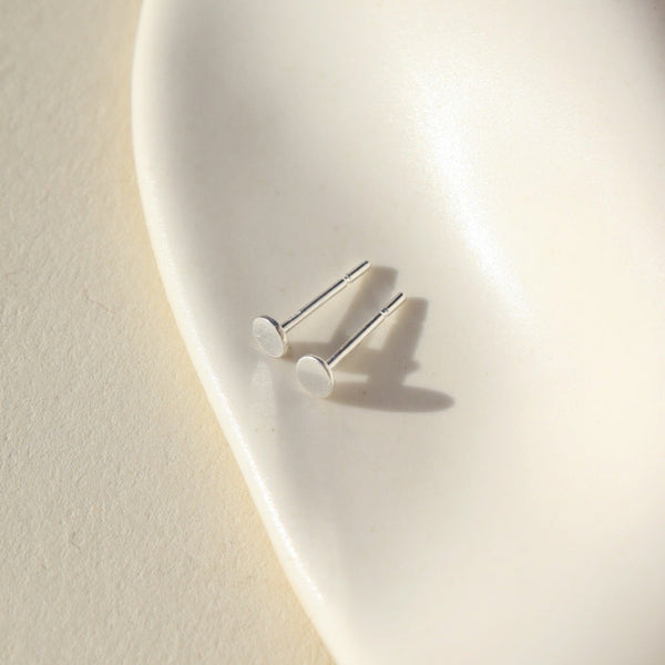 A pair of tiny silver dot shaped stud earrings on a neutral stone background.