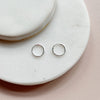 Two small silver hoop earrings resting on a white marble coaster against a soft pink background.