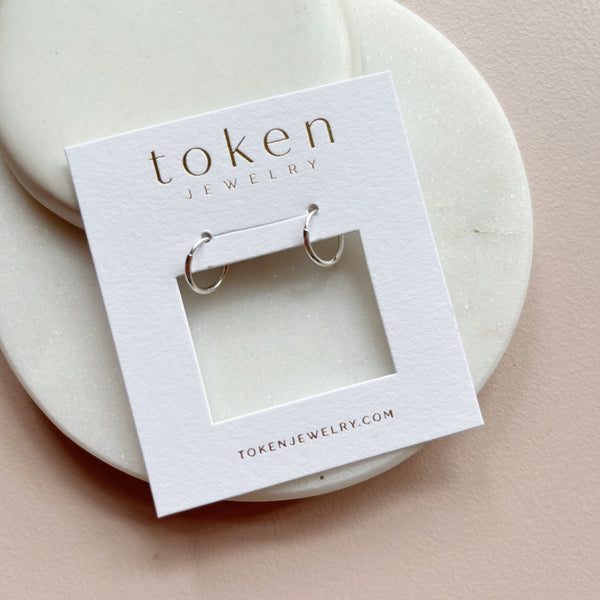 A pair of small silver hoop earrings displayed on a white jewelry card labeled 'token jewelry', placed on a round cream stand against a soft blush background.