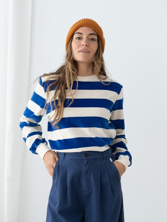 A woman stands indoors wearing a blue and white horizontally striped long-sleeve top, navy wide-leg pants, and an orange beanie. She is posed in front of a white curtain and wall.