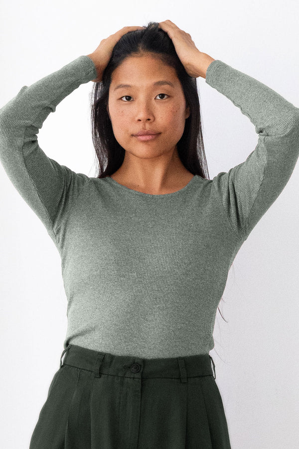 A woman with dark hair is wearing a heathered sage green long-sleeve knit top and dark green trousers, posing against a plain white background.
