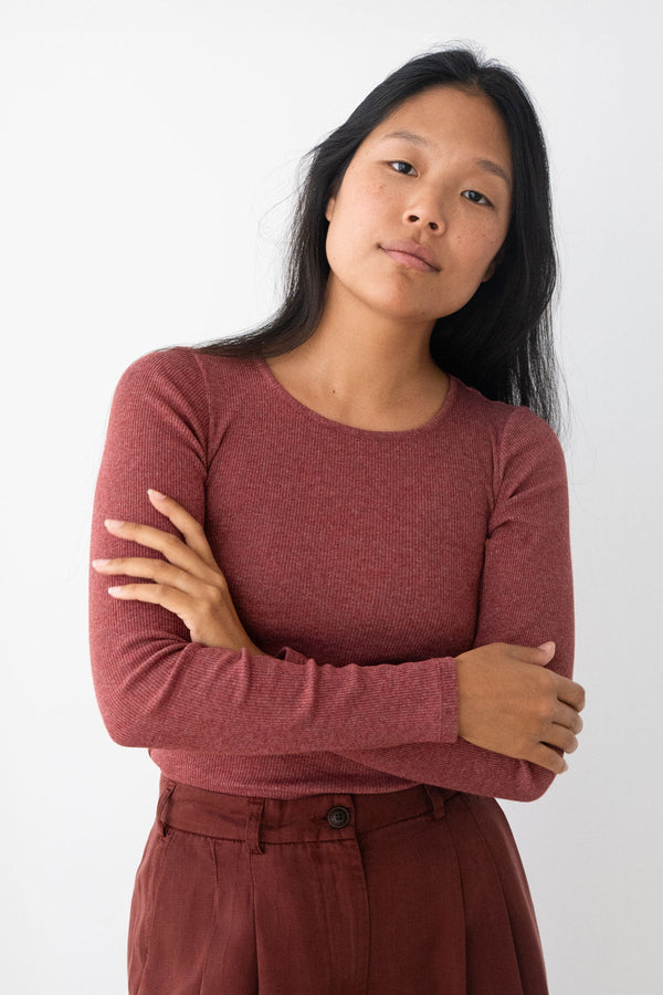 A woman with long black hair wearing a heathered dusty red long-sleeve crewneck top and matching dark red trousers, standing against a plain white background with her arms crossed.