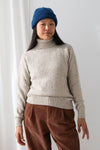 A person with long dark hair wearing a blue beanie, a beige turtleneck sweater, and brown corduroy pants standing against a white wall.