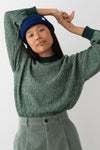 A model wearing a green and white striped knit sweater, a dark blue beanie, and sage green corduroy pants, posing against a light gray background.