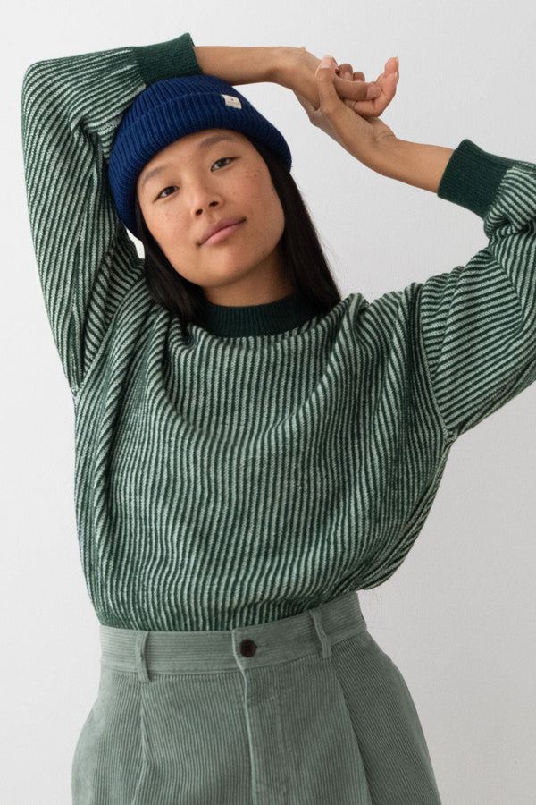 A model wearing a green and white striped knit sweater, a dark blue beanie, and sage green corduroy pants, posing against a light gray background.