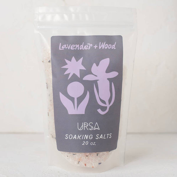 A clear stand-up pouch containing URSA Soaking Salts, featuring a dark gray label with lavender illustrations of a star, flower, and bird, and the scent name 'Lavender + Wood'.
