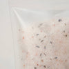 A close-up of a clear resealable pouch containing a light pinkish-beige bath salt blend with small scattered dark and muted orange flecks, photographed against a plain light background.