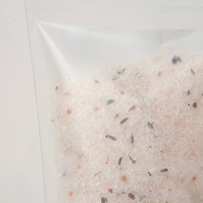 A close-up of a clear resealable pouch containing a light pinkish-beige bath salt blend with small scattered dark and muted orange flecks, photographed against a plain light background.