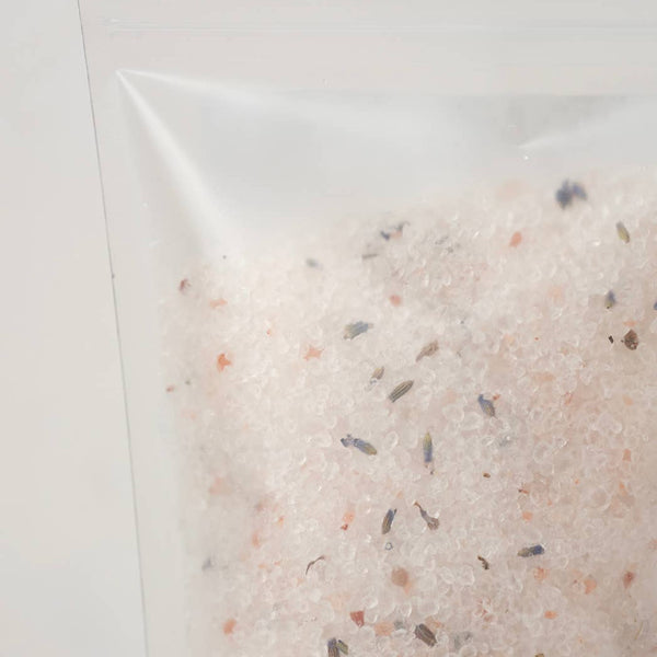A close-up of a clear resealable pouch containing a light pinkish-beige bath salt blend with small scattered dark and muted orange flecks, photographed against a plain light background.