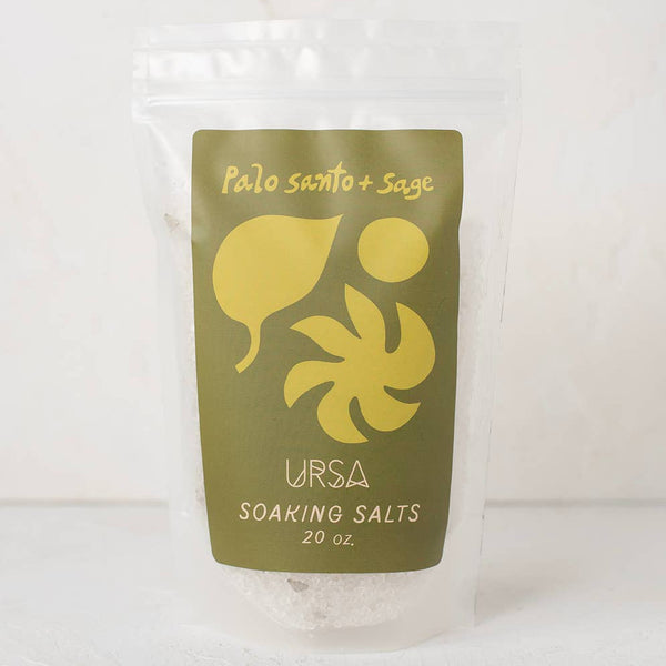 A clear stand-up pouch containing white soaking salts, with a large olive green front label reading 'Palo Santo + Sage' and 'URSA SOAKING SALTS 20 oz.' on a light neutral background.