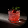 A red cocktail with a strawberry garnish in an Art Deco inspired lowball crystal tumbler against a dark surface and backdrop.