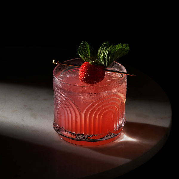 A red cocktail with a strawberry garnish in an Art Deco inspired lowball crystal tumbler against a dark surface and backdrop.