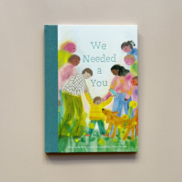 An illustrated children's book called 'We Needed A You' with a colorful design of a child surrounded by adults and other kids and a dog. The book sits against a neutral backdrop.