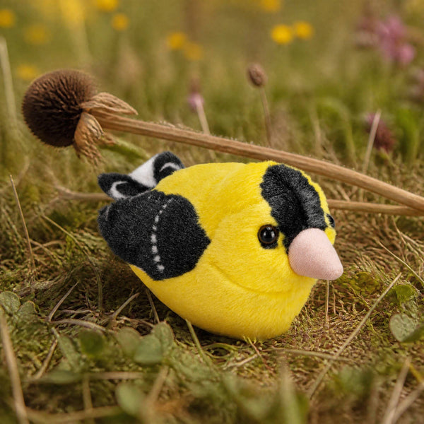 An American gold finch plush toy sits on a grassy wildflower surface.