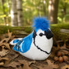 A plush toy blue jay with bright blue and white fur, black facial markings, and a fluffy blue crest, posed on forest floor leaves and moss with acorns and blurred trees in the background.