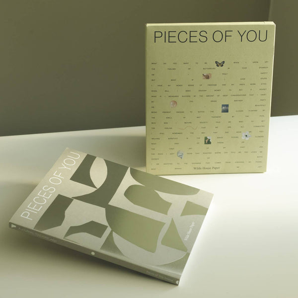 A guided journal called 'Pieces of You' fearing a geometric foil print cover, laid next to its paper packaging covered in text, photographed on a white surgface against a soft green wall.