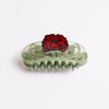 A clear acrylic hair claw clip featuring a light green scrollwork base and a prominent red poppy flower on top, photographed on a clean white background.