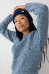 A person with long dark hair and a navy beanie wearing a blue marled knit wool sweater standing against a white wall.