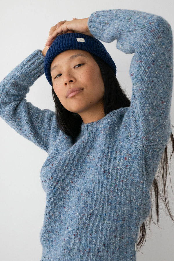 A person with long dark hair and a navy beanie wearing a blue marled knit wool sweater standing against a white wall.