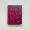 The party cookbook 'Your Turn to Host' by Amber Mayfield Hewett, featuring a deep magenta cover with bright pink text and illustrations, displayed against a neutral background.