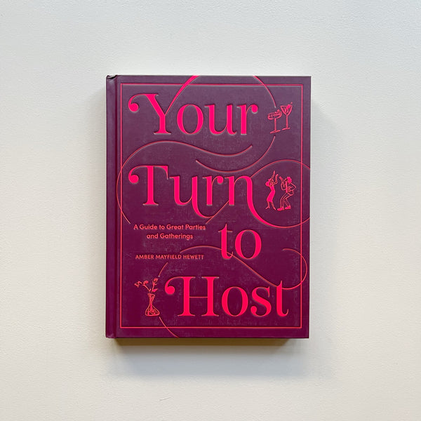 The party cookbook 'Your Turn to Host' by Amber Mayfield Hewett, featuring a deep magenta cover with bright pink text and illustrations, displayed against a neutral background.