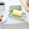 A pale green ceramic butter dish with a matching lid and a small metal spreader, shown with a stick of butter on a white tabletop. A cup of coffee, bread on a plate, and a wooden board are softly blurred in the background.