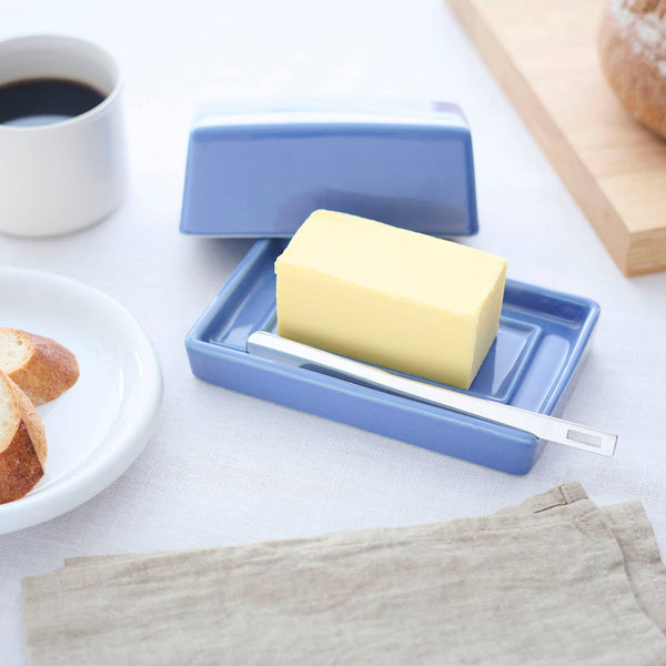A blue ceramic butter dish with a matching lid, holding a stick of yellow butter and a silver spreader, styled on a white tablecloth with a cup of coffee, bread, and a napkin nearby.