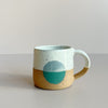 A clay espresso mug in natural clay and a dipped white speckle glaze. The natural and glazed clay are bisected by a teal green circle. The mug sits against a neutral backdrop.
