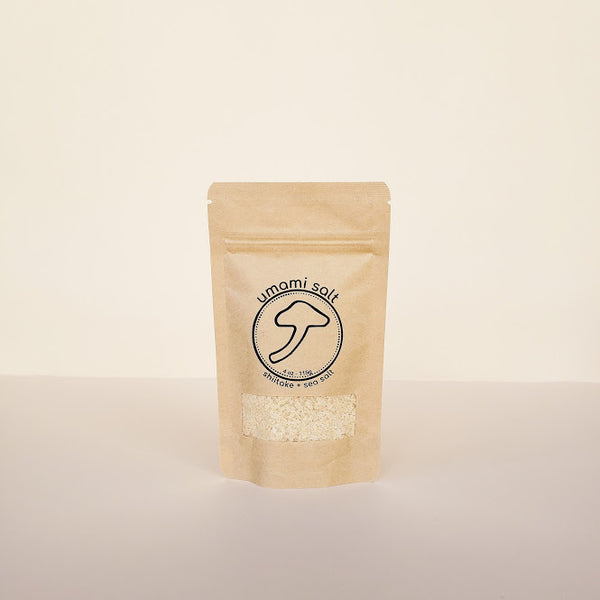 A standing pouch packaging of Umami Salt with a transparent window showing the product inside. The package is beige with a black label that includes the brand name and a mushroom illustration.
